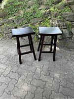 Two black glossy wooden bar stools outdoors on a paved surface with stone and moss background, showing overall condition and design.