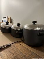 Three used black Circulon pots with silver lids, placed in a row. The lids have black knobs and the pots show signs of wear.