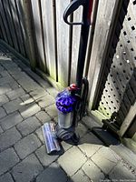 Dyson upright vacuum cleaner standing on stone pavement with motorized floor brush placed beside it, showing clear dustbin and purple cyclone.