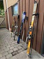 Four pairs of skis with bindings and four sets of ski poles leaning against a brown wall arranged on an outdoor stone floor.