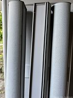 Close-up image of three roller shade tubes in white with soft grey fabric rolled around them, showing fabric texture and roller mechanism end caps.