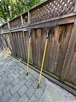 Two metal garden rakes with fan-shaped metal tines and yellow handles leaning against a wooden fence.