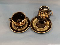 Two black ceramic cups and saucers with 24K gold decorative patterns arranged side by side, showing front and top views of the set.