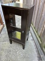 Full view of black stained solid pine cabinet with glass front door, standing outside on concrete surface.