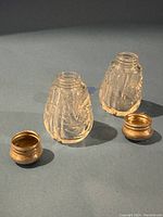 Two small vintage crystal salt and pepper shakers without tops alongside two metal caps on a grey surface.