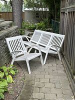 Outdoor white wooden couch and chair in weathered condition on stone patio against wooden fence and garden