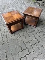Two wooden vintage side tables showing square tops and lower shelves from a side angle, placed on a paved surface.