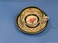 Tea cup and saucer set showing floral rose design inside cup and gold ornamental pattern on dark green background.