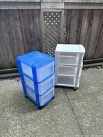 Two plastic rolling storage units pictured outside against a wooden fence, showing blue and clear frame units with three drawers each.