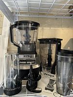 Photo showing Ninja Ultra Kitchen System with blender, food processor containers, blades, and smoothie cups all arranged on metal shelf.