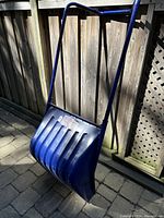 Blue plastic push snow shovel shown standing on patio ground against wooden fence, showing full handle and scoop shape
