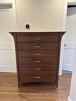 Tall wooden dresser with six drawers, two bottom drawers deeper than the top four, medium wood tone, modern metal handles, placed on hardwood floor against a light-colored wall with white door frames.