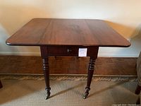 Front view of the antique wood drop leaf table with leaves open, showing the drawer and turned legs on castors.