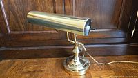 Side view of brass desk lamp showing cylindrical adjustable shade and round base with power cord.