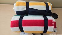 Two rolled and strapped Hudson Bay fleece blankets, one on top of the other, showing iconic multi-color stripes in yellow, white, red, green, and navy on cream background.