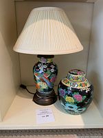 Photo taken of the lamp and the urn placed adjacent on a white shelf, showing the urn's bright floral design and the lamp's pleated cream shade.