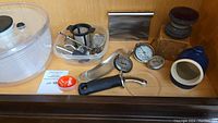 Photo showing OXO salad spinner, metal herb cutter, handheld grater, metal thermometer, blue ceramic garlic keeper, tea strainers, pizza cutter, and smaller containers.