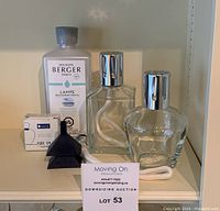 Photo showing both glass oil lamp burners, Ocean Breeze refill bottle, wick with protective cap, and boxed catalytic burner arranged on a shelf with a business card in foreground.