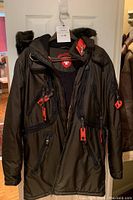 Front view of black Wellensteyn parka with red zipper pulls and faux fur-trimmed hood hanging on door