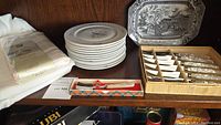 Full view of shelf with plates stacked, tablecloth and napkins folded, boxed steak cutters and serving spoon visible
