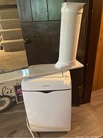Front view of white portable Noma air conditioner with attached flat vent panel and flexible vent hose extending upward.
