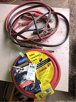 Photo of coiled red rubber air hose in original packaging labeled Colorite alongside used booster cables with red and black clamps on a table.