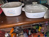 Two vintage Corningware casserole dishes with floral Blue Basket pattern on a wooden surface, each with a clear glass lid.