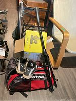Pitching machine in box with tripod next to Mizuno baseball shoes on Easton sports bag