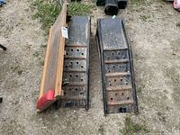 Photo displaying two black metal car ramps with holes and one wooden mechanic dolly with red tips on casters placed on the ground outdoors.