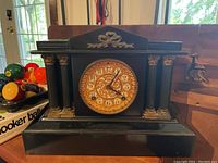 Front view of vintage heavy mantel clock showing the ornate dial, black body and decorative columns.