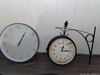 Two wall clocks side by side on floor against wall. One is a white-faced single-sided clock 14 inches diameter. The other is a Louis Barnard two-sided metal black framed clock 10 inches diameter with mounting bracket.