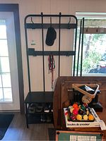 Full view of the black brushed metal coat rack with bench and shoe storage shelves below, showing hanging hooks with a hat and leashes, shoes on the shelves, located near a door and window.