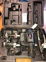 Open black case showing Dremel rotary tool and multiple attachments including flexible shaft, clamp mount, and bits.
