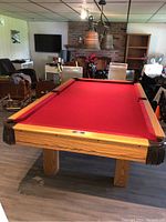 Front angled view of Dufferin pool table showing red felt surface, oak wood rails with six pocket openings and brown leather corner pockets with black fringe nets.