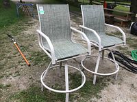 Two lawn chairs with white metal frames, circular footrests, armrests, and grey-green mesh fabric seats and backrests, placed outdoors on grass and gravel.