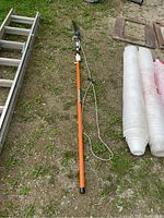 Full view of the orange Stihl pole saw laying on grass with saw head on one end and pull cord on the other.