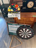 Pool balls and snooker balls box on wooden surface, clock in background, pool table game chart and dartboard leaning against furniture, wooden pool triangle rack on floor