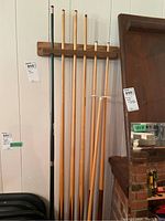 Six wooden pool cues and a bridge stick in a wooden wall-mounted rack against a light-colored wall.