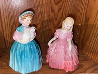 Two Royal Doulton figurines on wooden shelf: girl in blue dress holding bunny and girl in pink dress holding rose.