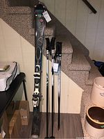 Full view of skis and two sets of poles leaning against stairs indoors, showing condition and size tag.