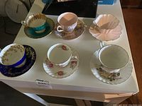 Overview of all six teacups with matching saucers on white table showing different styles and colors.