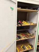 Storage cabinet showing multiple shelves with assorted hand tools, dowels, safety glasses, and a respirator. Tools include files, punches, box cutters, and a rasp.