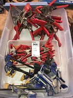 Overhead view of multiple red-handled spring clamps and blue Irwin Quick-Grip bar clamps inside a plastic bin