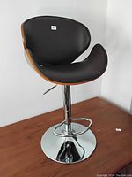 Front and side view of the adjustable stool showing black cushioned seat, curved wooden back and chrome base with footrest and adjustment lever.