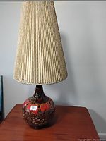 Front and slight side view of ceramic table lamp showing textured rounded base with red, brown, black glaze and tall pleated beige shade.