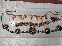 Four copper jewelry pieces laid out on white background: two necklaces, one bracelet, one brooch, showing shapes and details.
