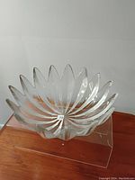 Clear large crystal bowl with petal-shaped edges and radial pattern inside, photographed on a wooden surface with white wall background.