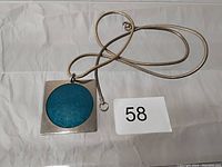 Top view of the silver-tone snake chain necklace with a square pendant with blue circular enamel inlay, tagged with '58'.