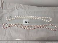 Two pearl necklaces displayed on a soft surface with a lot number 63 tag. White pearls necklace has a decorative clasp.