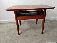 Front-side view showing the overall teak construction, four round legs, and a lower shelf with black surface.